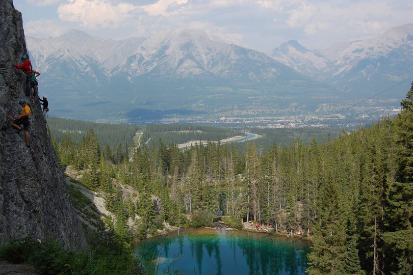 Several people are rock climbing on a cliff overlooking a valley with a river and forest. In the background, there are majestic mountains.