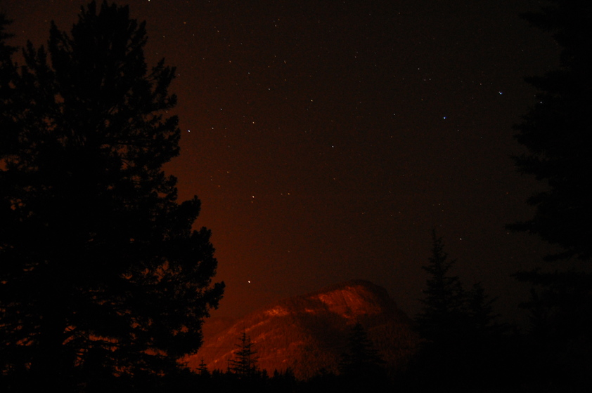 A dark night sky with silhouettes of trees in the foreground and a mountain illuminated by orange light in the background. Stars are visible scattered across the sky.