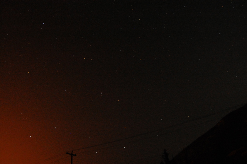 Night sky with stars visible against a dark background, with faint orange glow from city lights at the bottom.