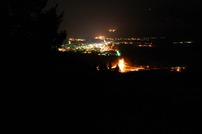 A nighttime view of Banff from a hilltop, with illuminated streets and buildings creating a glowing effect against the dark sky. Trees in the foreground frame the scene.