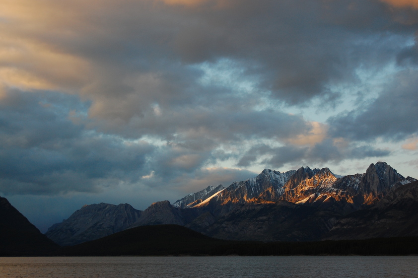 A peaceful scene of a lake at the foot of a rugged mountain range, with clouds drifting across the sky.
