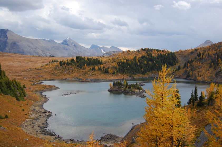 A scenic view of a lake nestled in the mountains with colorful trees in the foreground on a cloudy day.