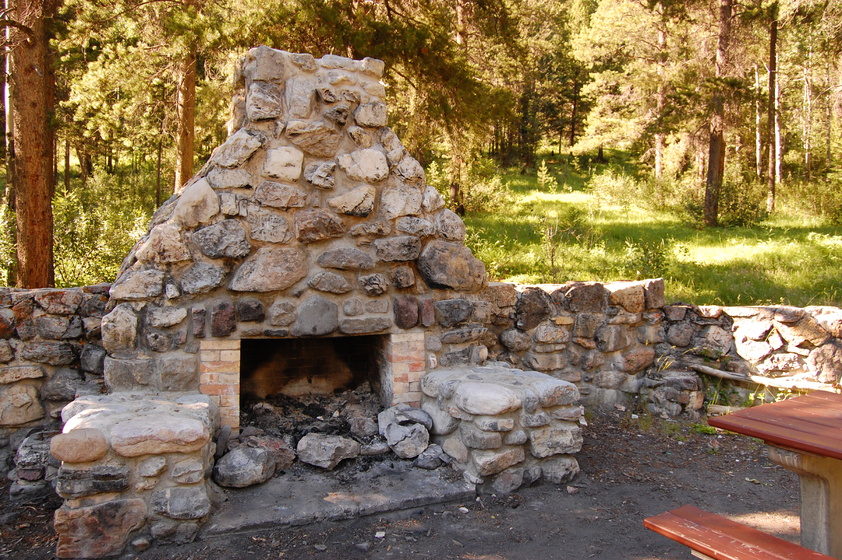 A rustic stone fireplace and seating area in a forest clearing, surrounded by trees and with remnants of a recent fire in the fireplace.
