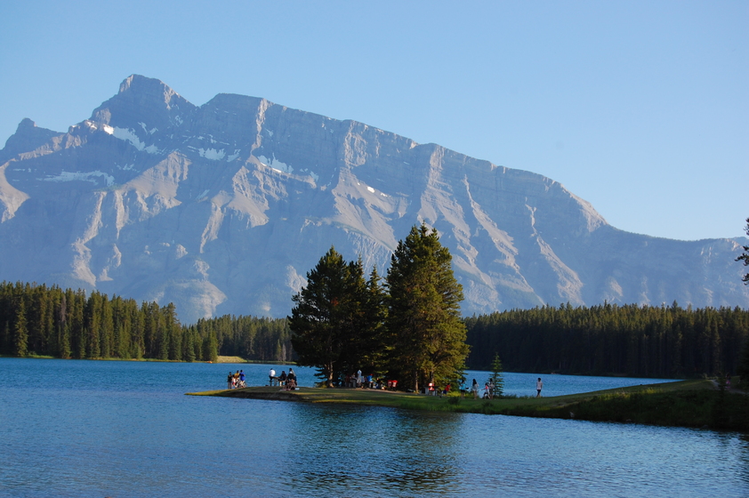 A serene lake surrounded by tall trees with a small peninsula in the center hosting a group of people enjoying outdoor activities, backed by majestic mountains.