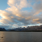 A calm lake at sunset, reflecting the colorful sky and silhouetted by mountains in the distance.