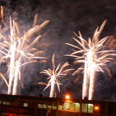 A white fireworks display erupts in a black night sky, with the silhouette of a building in the foreground.