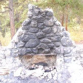 A stone fireplace surrounded by trees in a forest, with ashes and burnt wood in the fire pit.