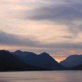 A serene lake nestled amidst towering mountains at dusk, with pastel-colored clouds filling the sky.