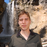 A man standing in front of a waterfall in a rocky outdoor environment.